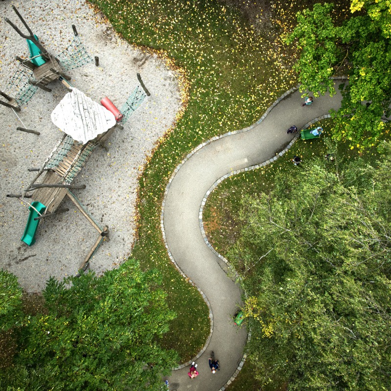 Aufnahme aus der Vogelperspektive, die einen Spielplatz mit vorbeischlängelndem Weg zeigt. Er ist eingebettet in eine grüne Umgebung. 