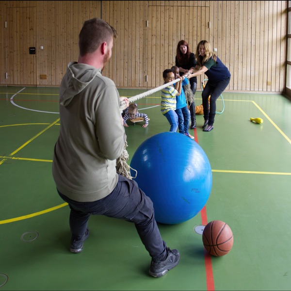 Personen spielen Seilziehen. Im Vordergrund ist eine Person, die alleine am Seil zieht. Auf der gegenüberliegenden Seite sind zwei Erwachsene und zwei Kinder, die gegen die andere Person antreten. Alle Personen befinden sich in einer Sporthalle. 