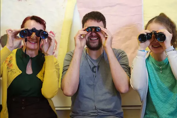 Drei Personen in unterschiedlicher, bunter Kleidung schauen jeweils mit einem Fernglas in Richtung Kamera. 